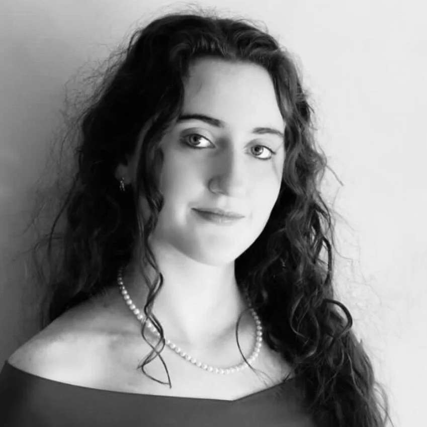 Black and white portrait of a young woman with curly hair, wearing a pearl necklace and earrings, looking at the camera with a slight smile, against a plain background.