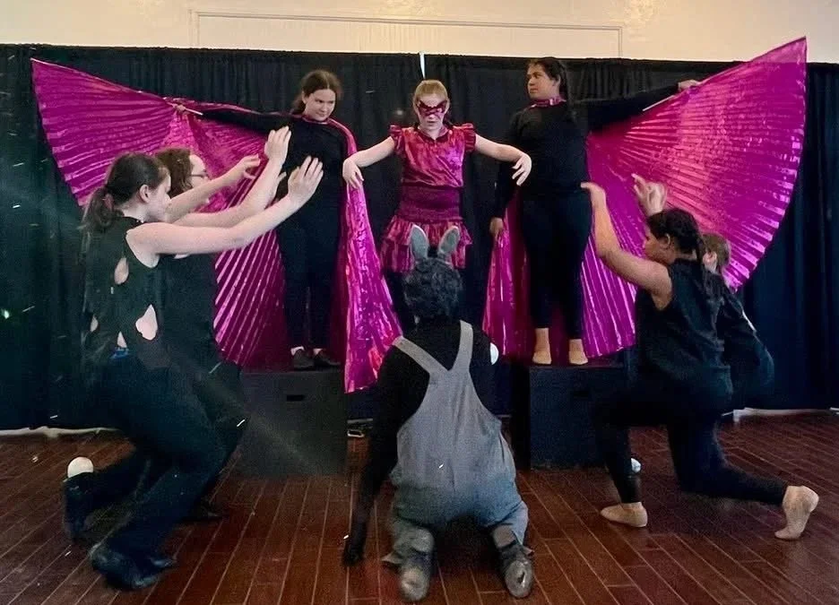 Children performing a play on stage with a purple butterfly wing backdrop, dressed in costumes including a girl in pink with face paint and a person in a bunny costume.