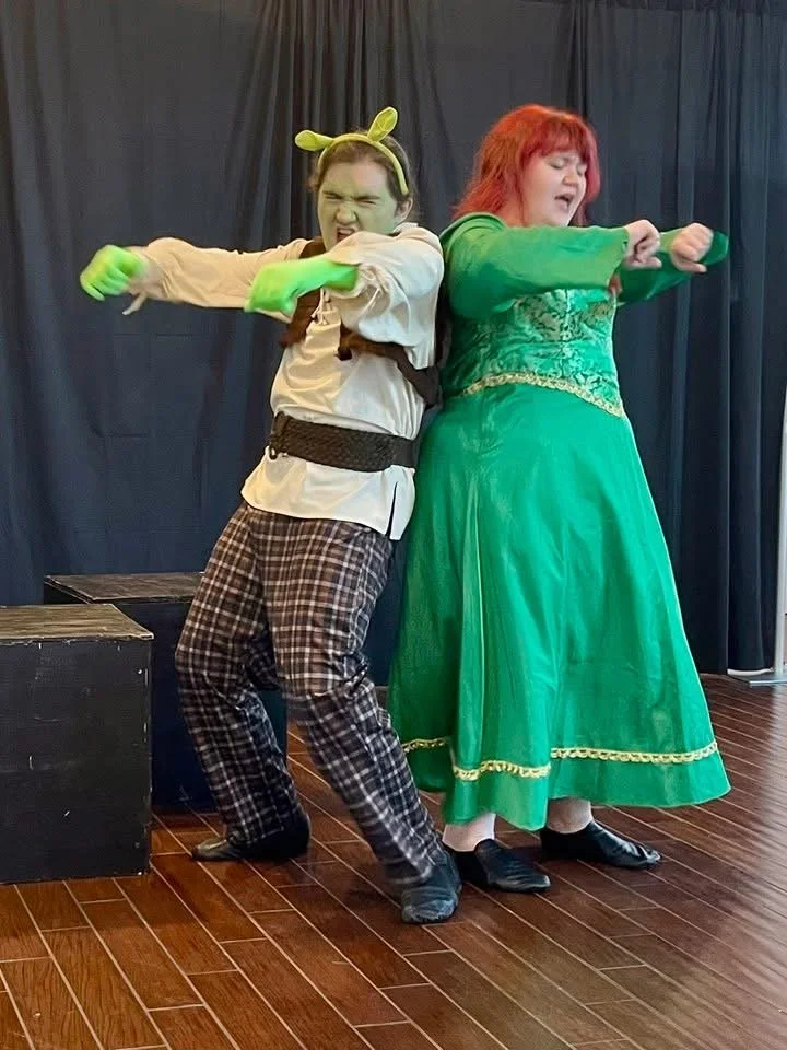 Two performers in colorful costumes and exaggerated expressions, dancing or acting on a stage with dark curtains. The performer on the left is dressed as a playful character with green face paint, a headband with ears, and plaid pants, while the one on the right wears a green dress with gold accents and has red hair.