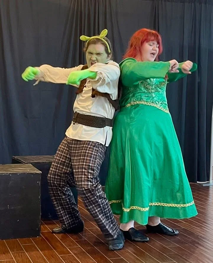 Two women in costumes dancing or performing on stage, one dressed as Shrek and the other in a green dress with orange hair.