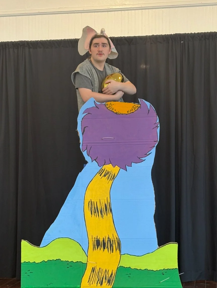 Person performing on a puppet stage with a colorful cat puppet and a headpiece in front of dark curtains.