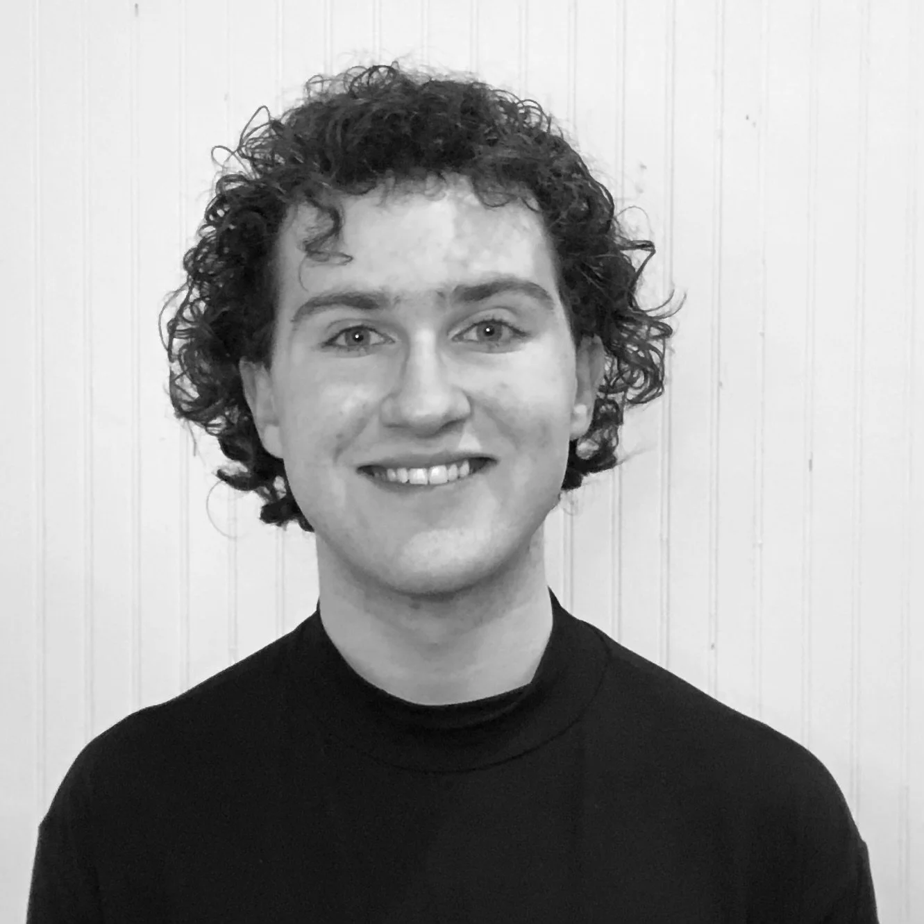 Black and white photo of a young person with curly hair smiling, wearing a black turtleneck top, standing in front of a light-colored wooden wall.