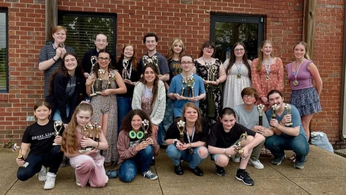 A group of young people posing outdoors in front of a brick building, holding trophies, celebrating an achievement.
