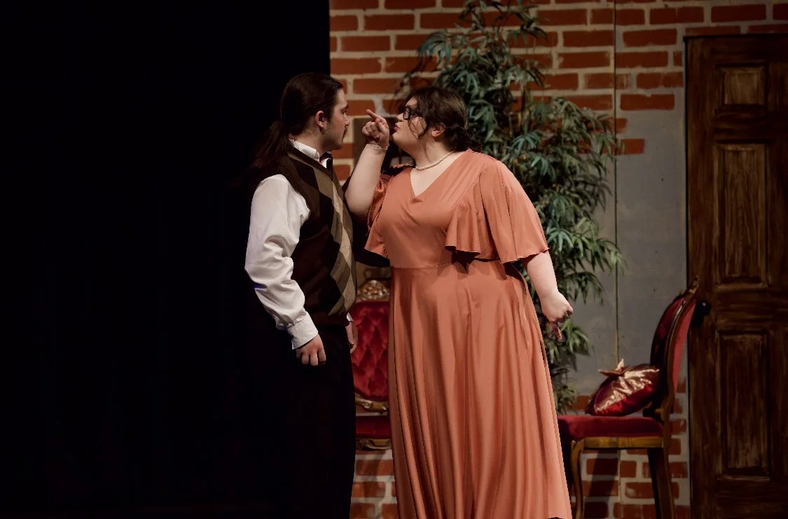 Two actors performing on stage, with one person in a white shirt and vest, and the other in an orange dress, engaging in a dramatic scene.