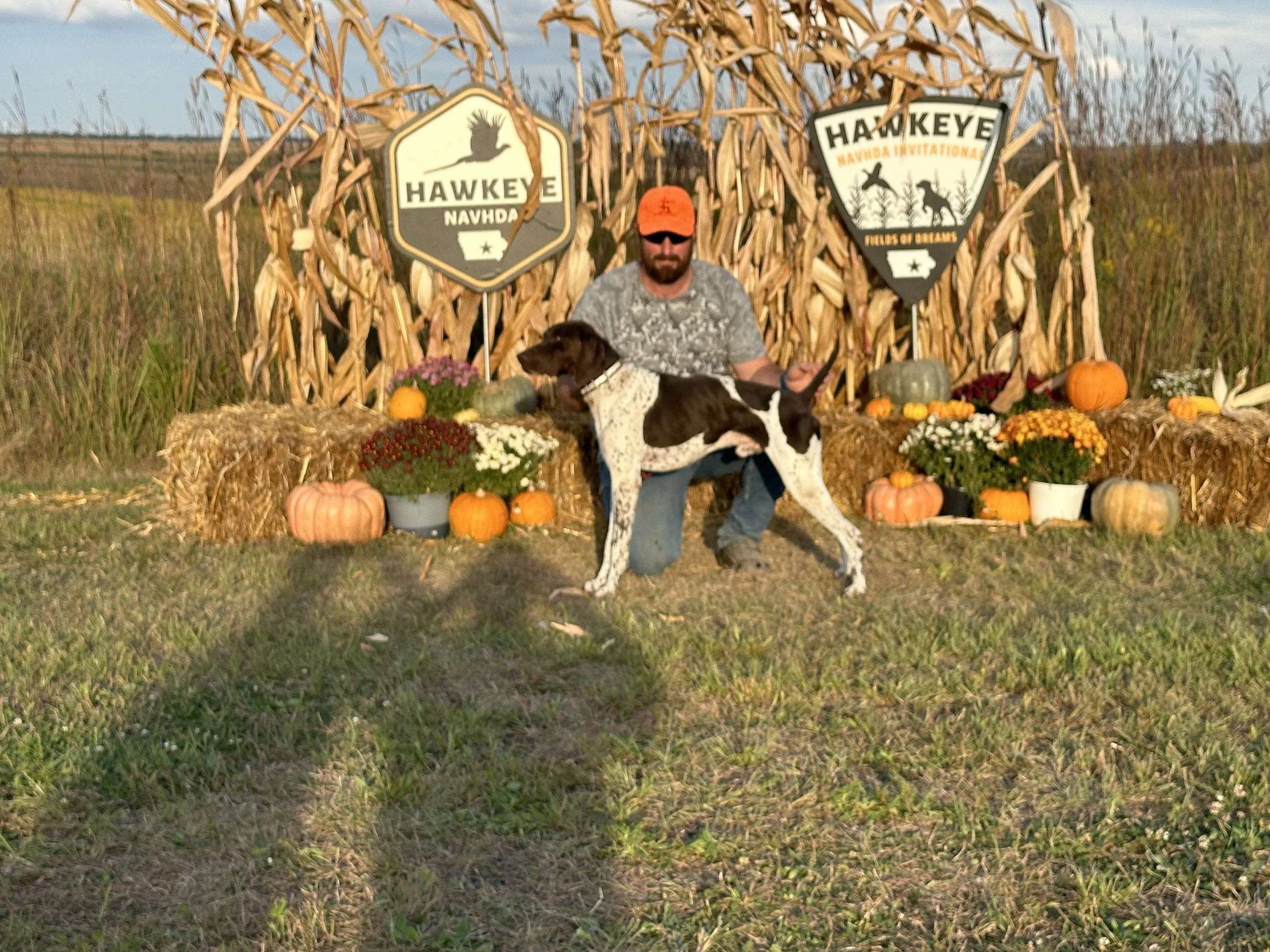 A man kneeling with a dog in front of fall harvest decorations, including pumpkins, gourds, hay bales, chrysanthemums, and autumn leaves, with signs reading 'Hawkeye Navhda' and 'Fields of Dreams' in the background.