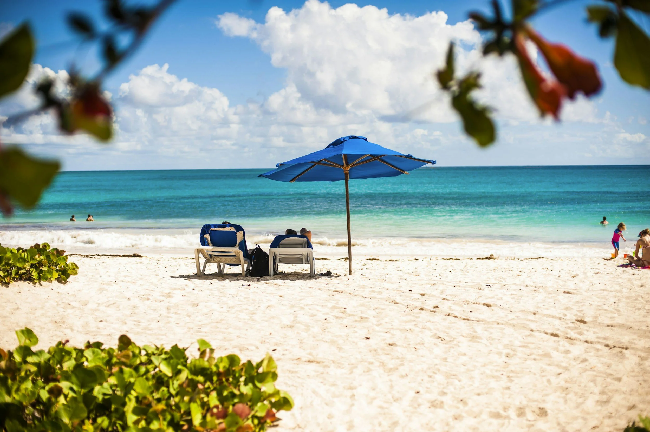 Beach scene with a blue umbrella, two people sitting on lounge chairs, children playing, and the ocean in the background under a partly cloudy sky.