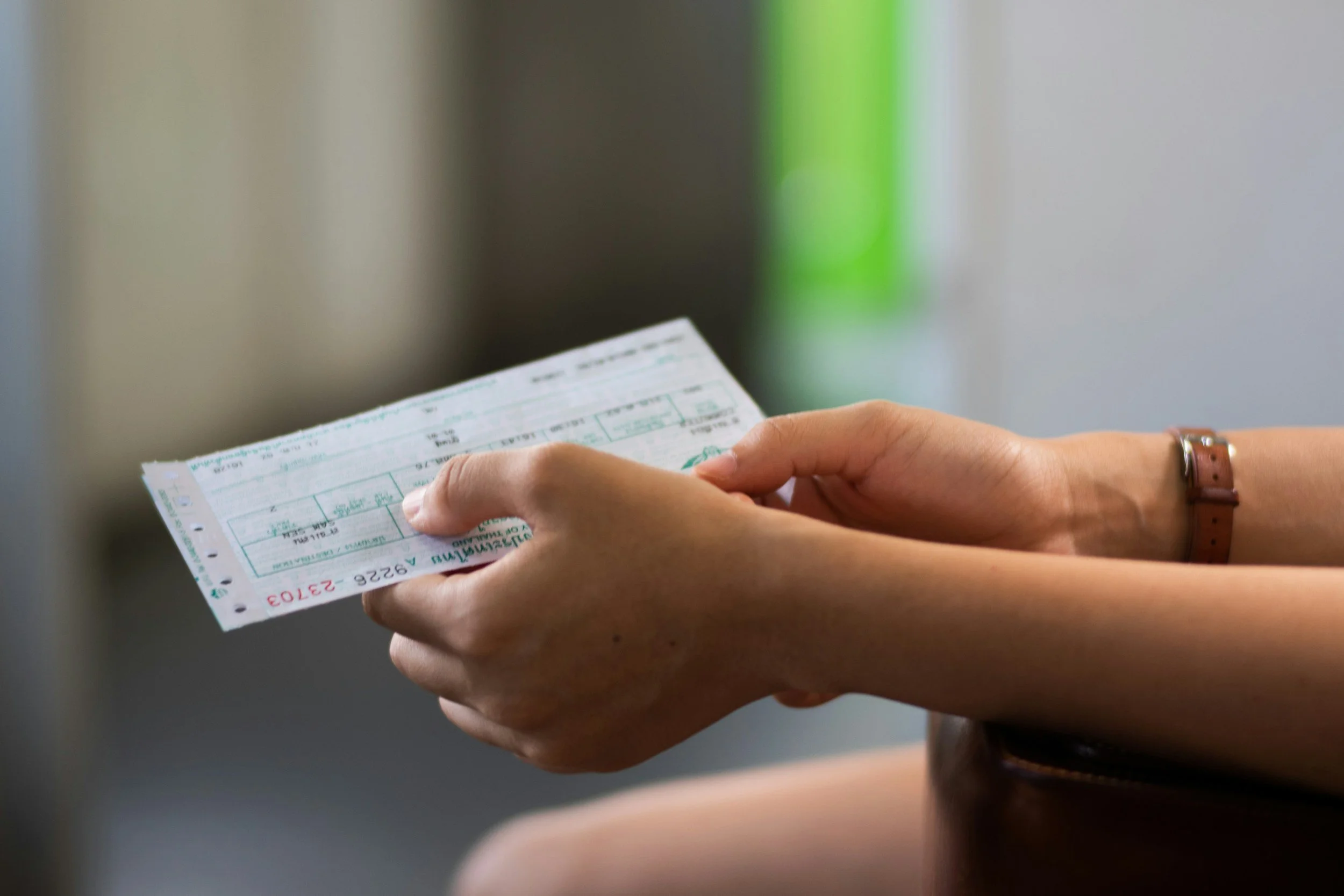 Person holding a boarding pass or ticket in hand, wearing a brown wristwatch, with a blurred background.