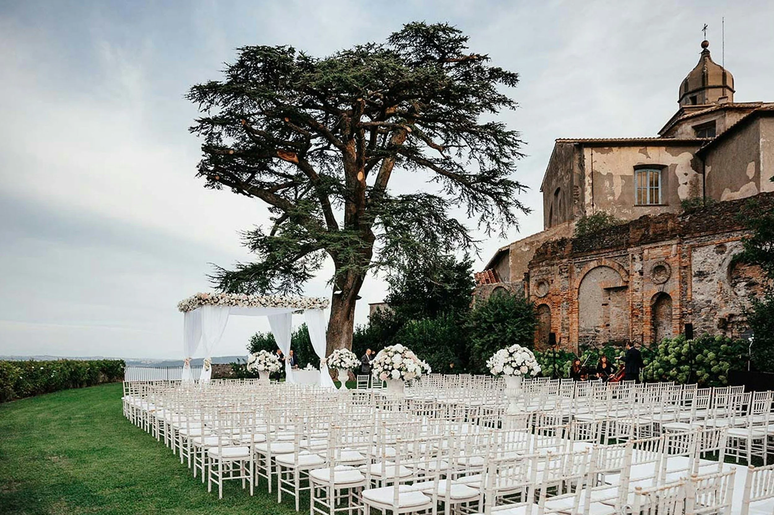 Outdoor wedding setup with white chairs and floral arrangements beneath a large tree, near an old stone building with a dome.