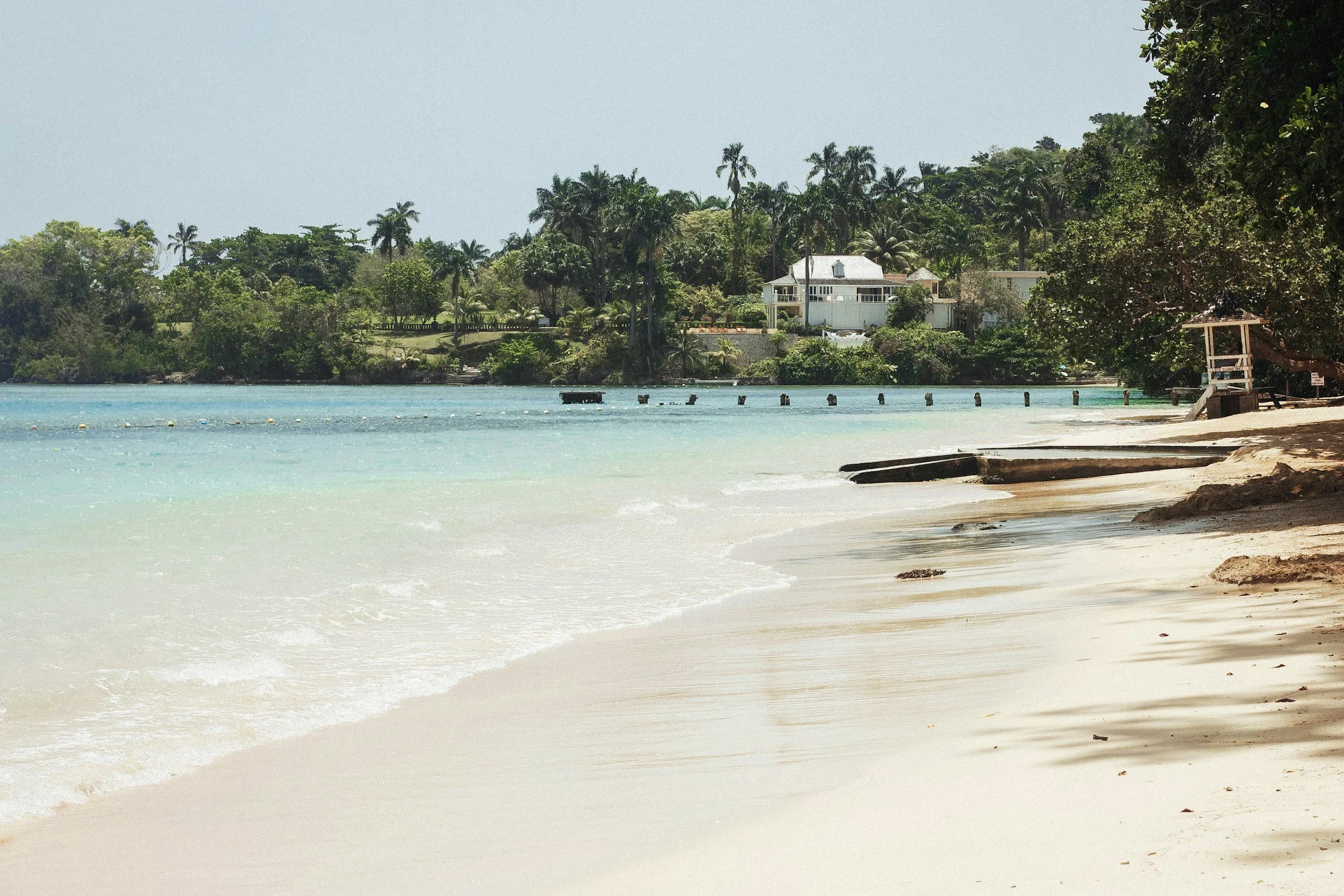 Beach with white sand and turquoise water, green trees and houses in the background.