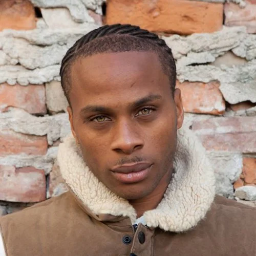 A young man with braids and a serious expression, standing in front of a brick wall