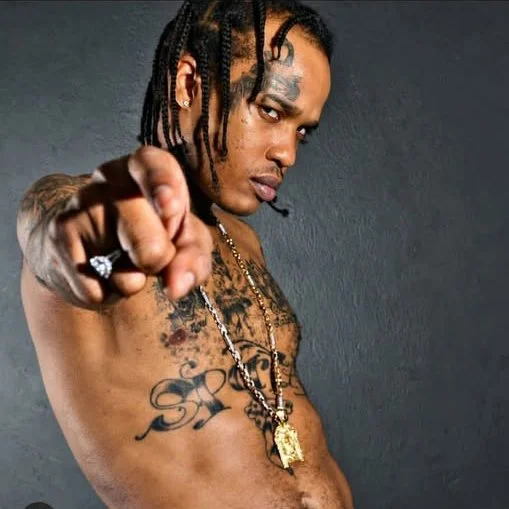 A shirtless man with tattoos and braided hair pointing directly at the camera against a dark background.