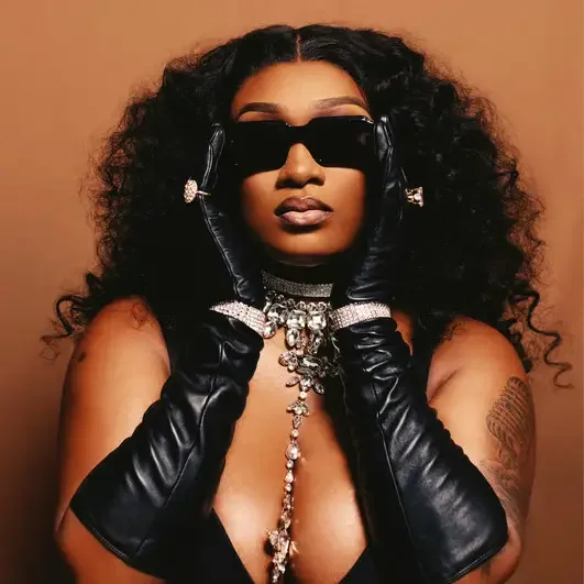 A woman with long curly hair wearing sunglasses, black leather gloves, and jewelry, striking a pose against a neutral background.