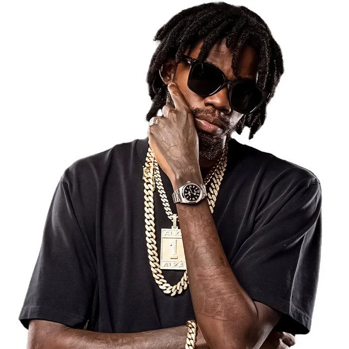 A man with dreadlocks wearing sunglasses, a black t-shirt, gold chains, a wristwatch, and a tag on his shirt, posing with his hand near his face against a white background.