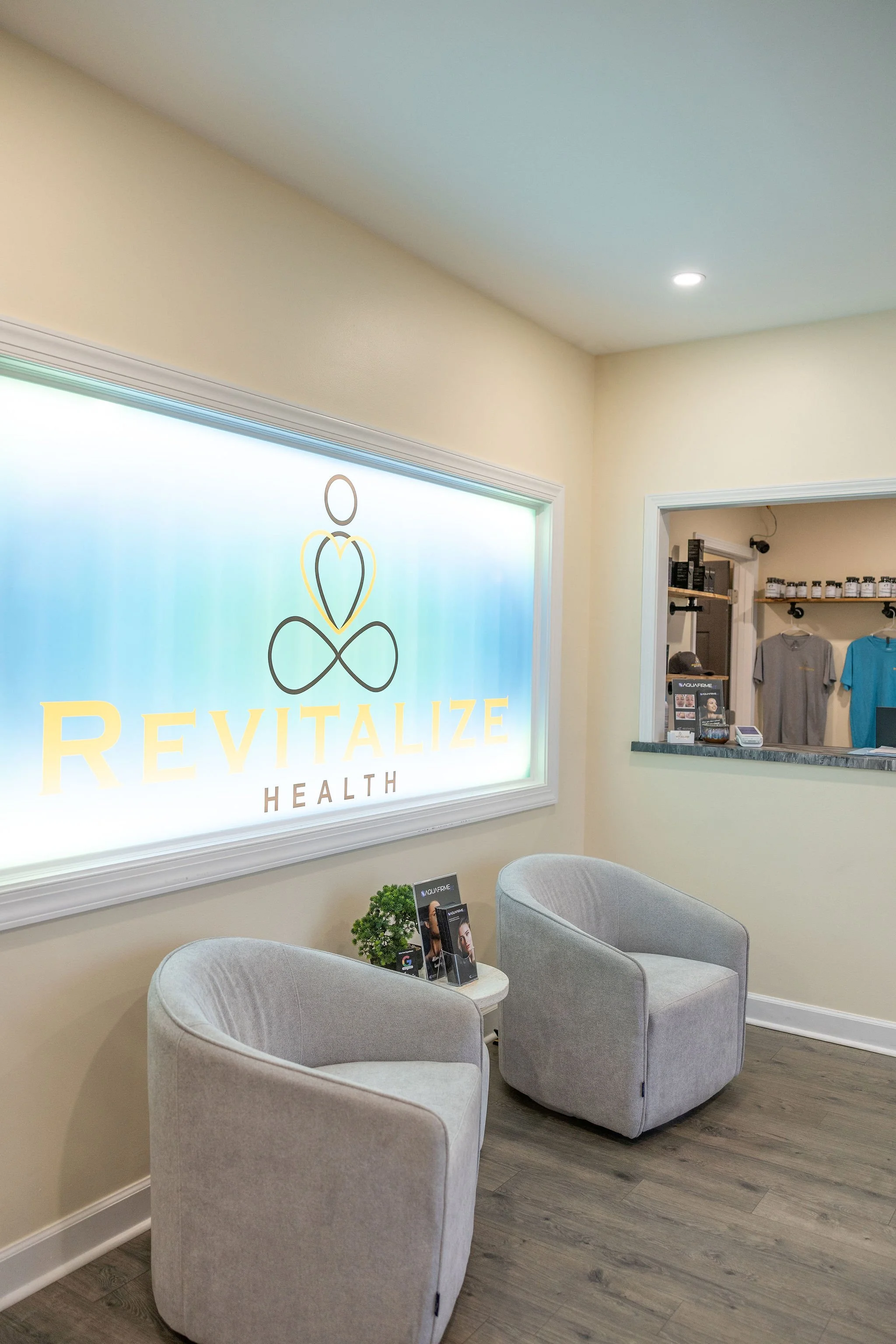 Waiting area with two gray armchairs, a small round table with a plant and magazines, a backlit sign reading 'REVITALIZE HEALTH,' a reception window, and a wall with retail products and clothing.