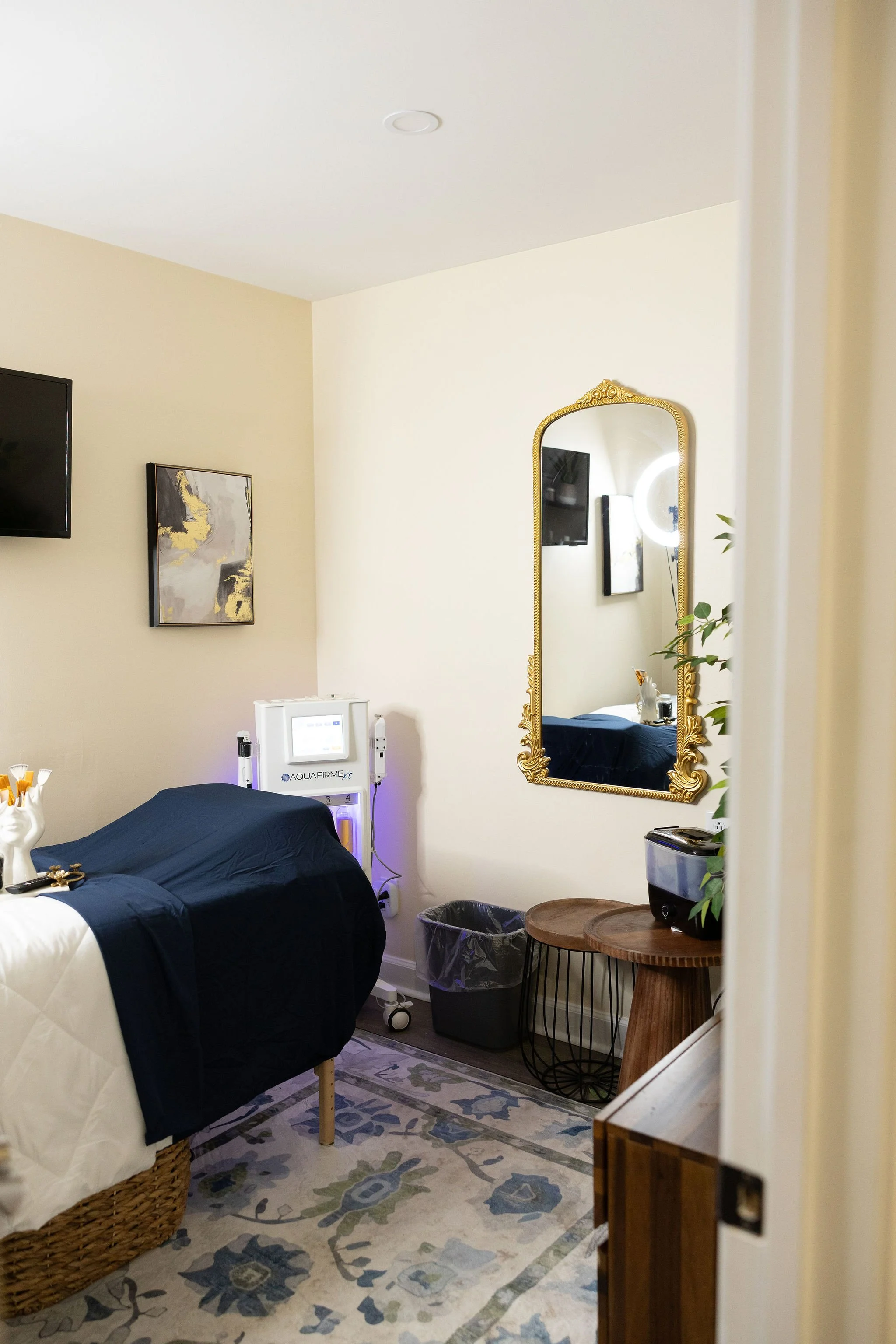 A treatment room with a bed covered in dark blue sheets, a decorative woven basket under the bed, a patterned rug, a small round wooden table, a trash can, a large framed mirror with an ornate gold frame, a wall-mounted TV, a piece of abstract artwork, a painting, and a small medical device labeled 'AQUAFIRME' in the corner