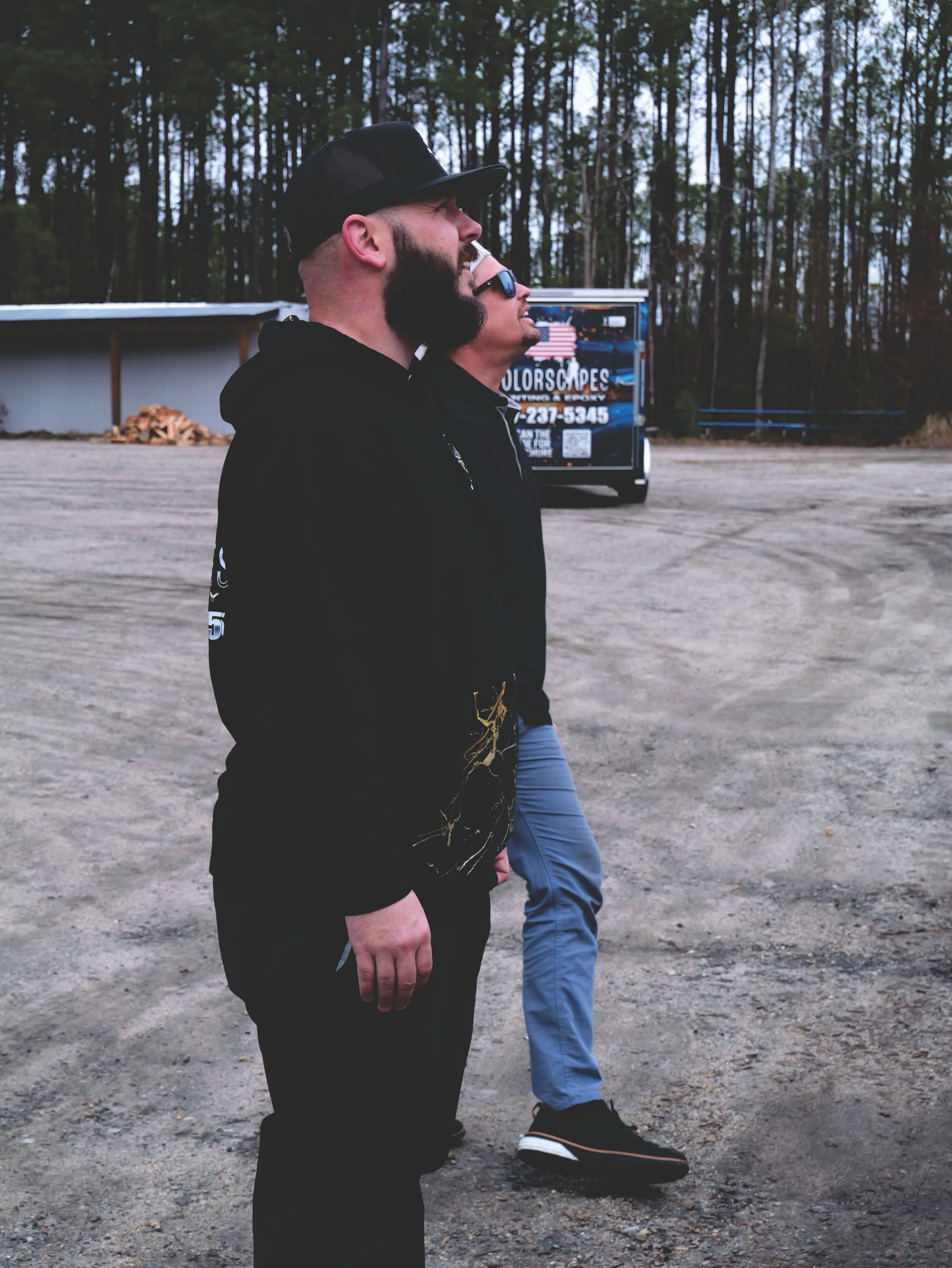 Two men standing outdoors on a dirt surface, one wearing a black hoodie and cap, the other wearing sunglasses and a black jacket, facing to the right. A truck with signage is visible in the background, along with a wooded area.