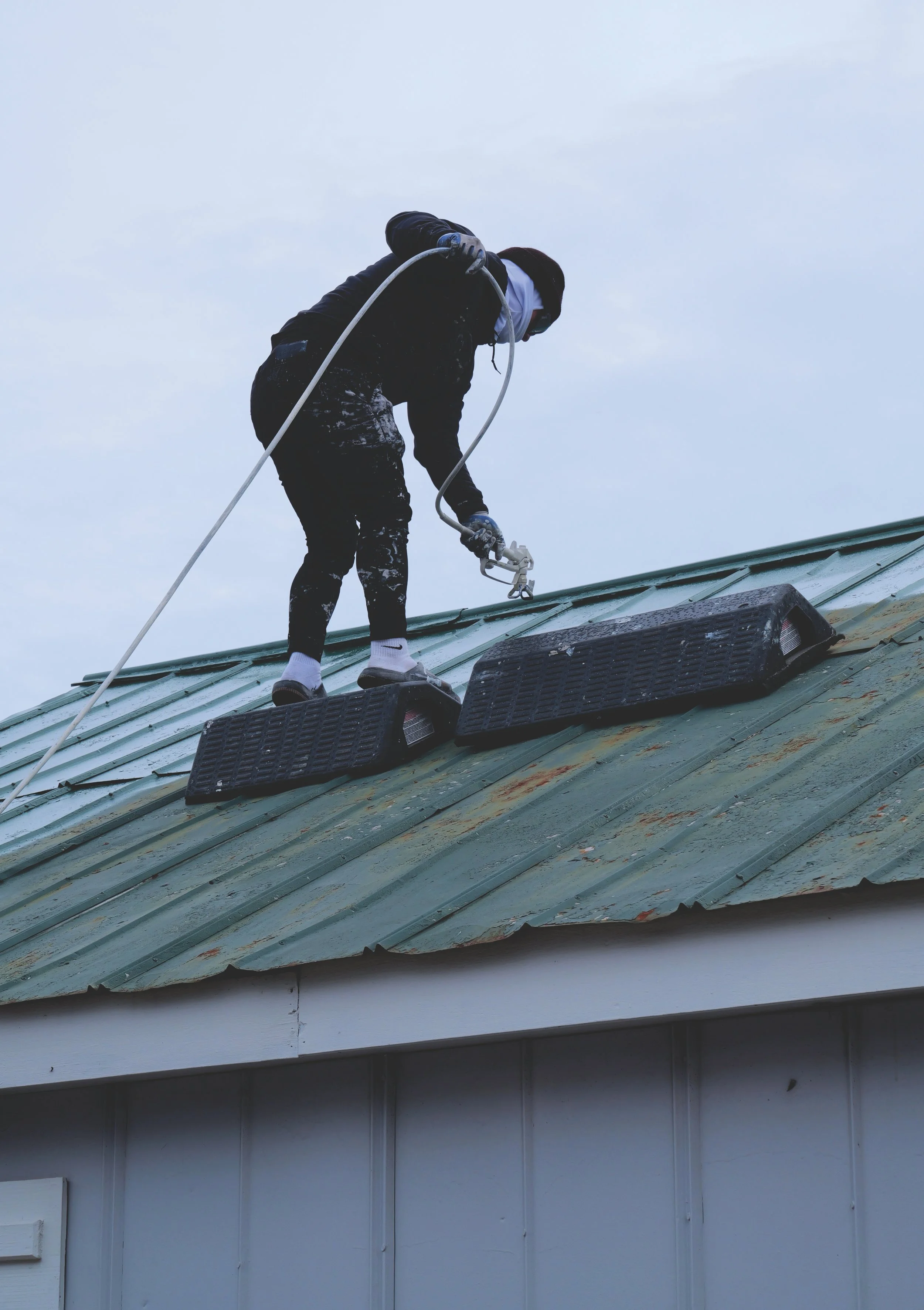 A person cleaning a roof with a spray device, standing on rubber mats. The roof is metal with rust stains, and the person is wearing protective gear including gloves and a face mask.