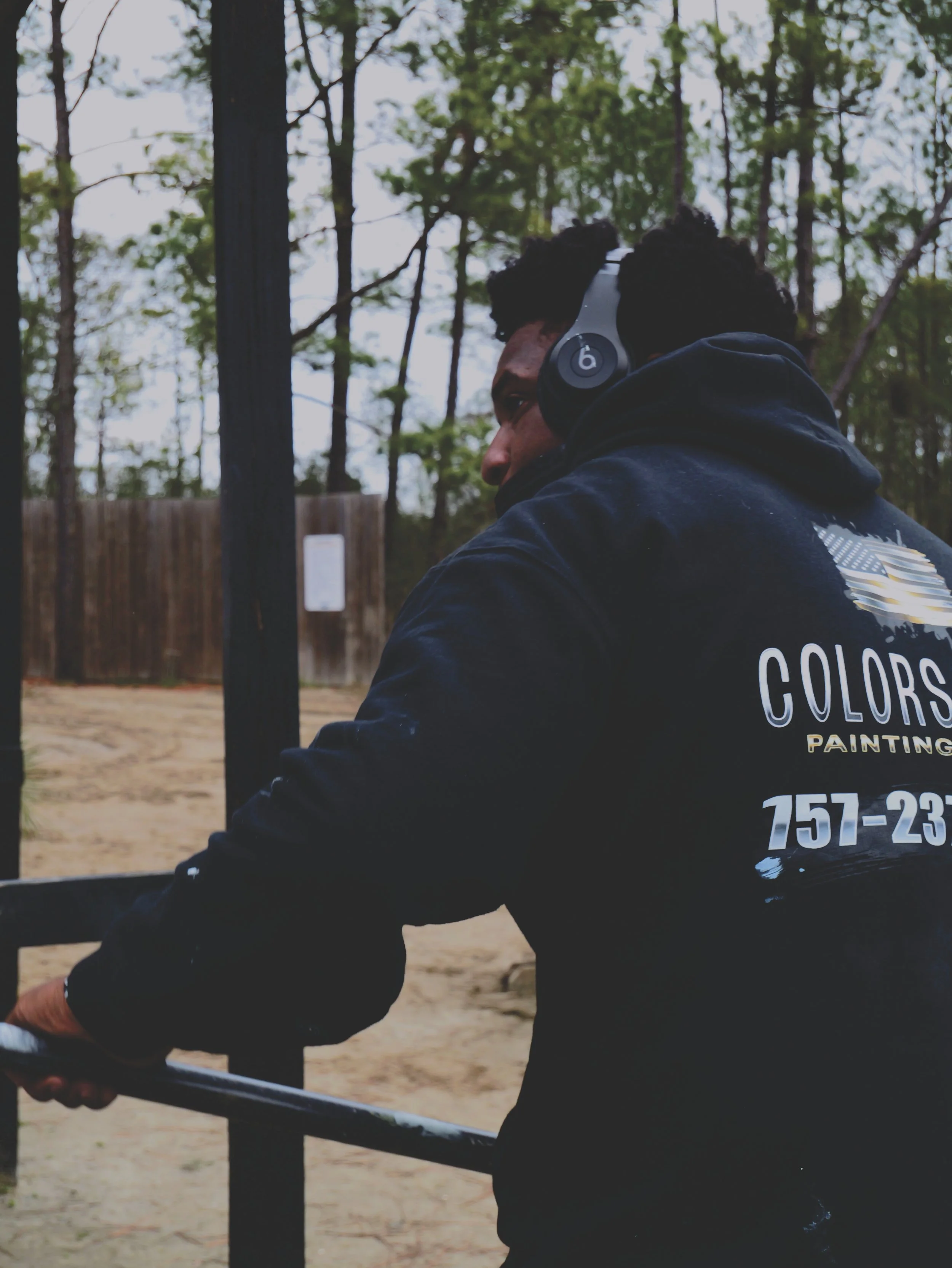 A man with curly hair wearing black headphones and a black hoodie with the text 'COLORS PAINTING' and a phone number on the back, holding a paint roller outdoors, near trees and a wooden fence.