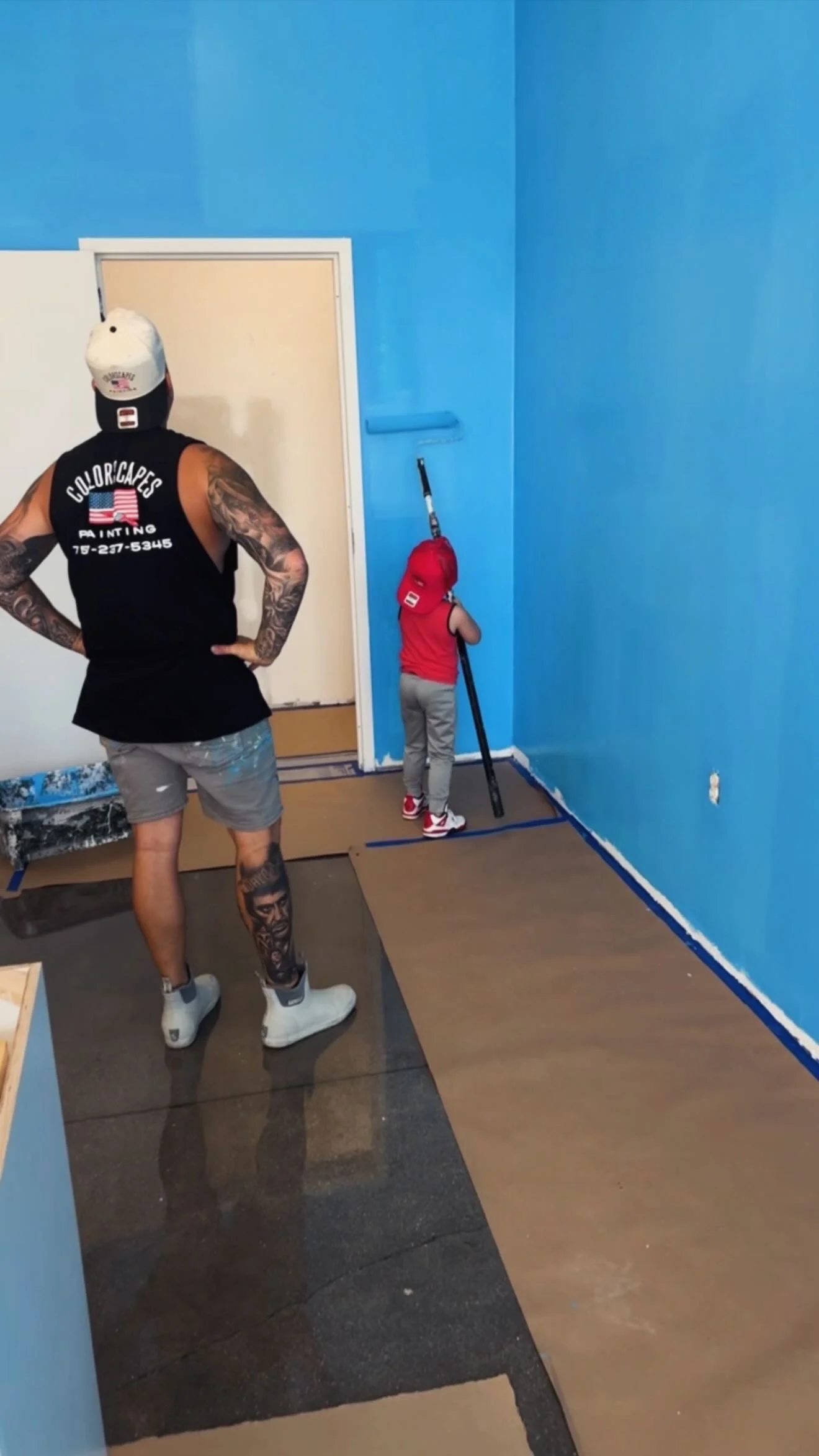 A man and a young boy painting a room blue. The man stands with hands on hips, wearing a black tank top, gray shorts, white sneakers, a white cap, and has tattoos. The boy is facing the wall, wearing a red sleeveless shirt, gray pants, red and white sneakers, and a red cap, holding a paint roller, painting the wall blue.