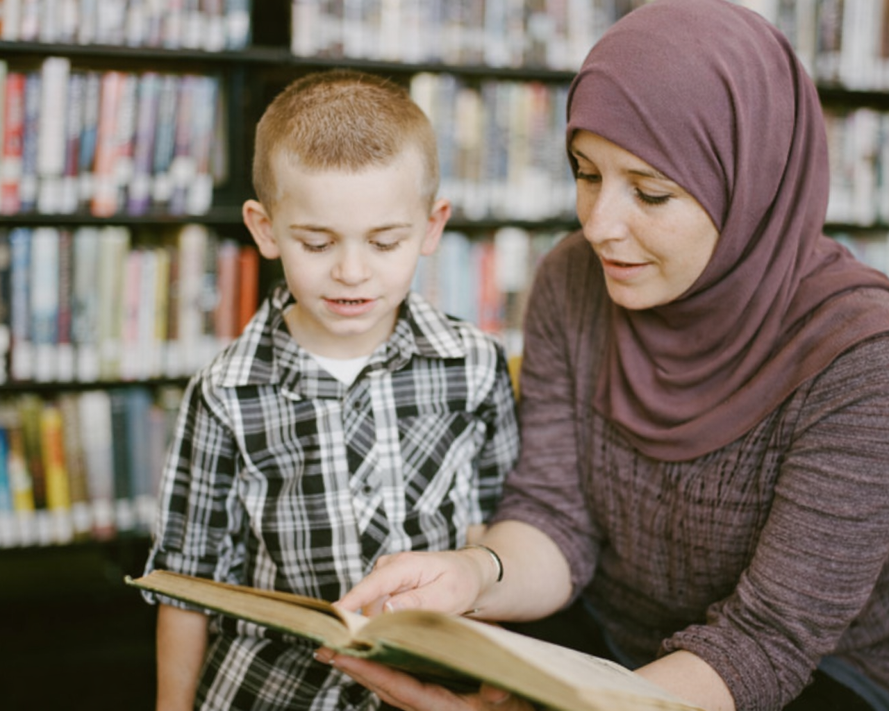 A young boy and a woman with a hijab reading a book together in a library.
