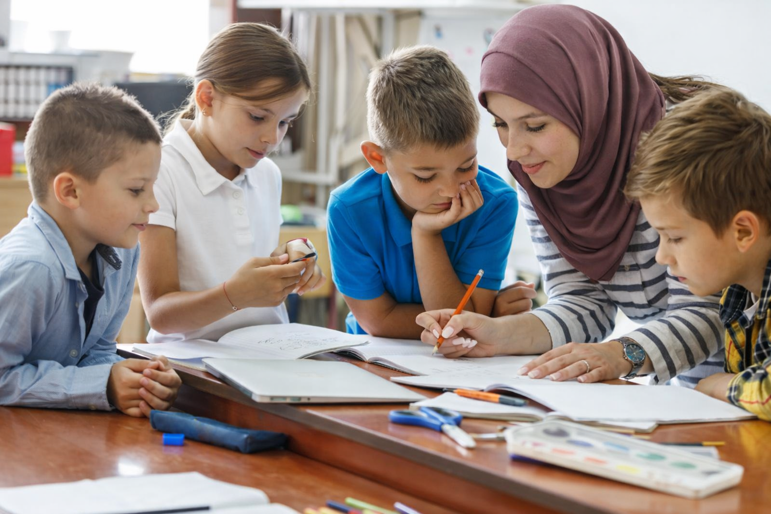 A female teacher shows and explains something in a book to four children gathered around a table during class.