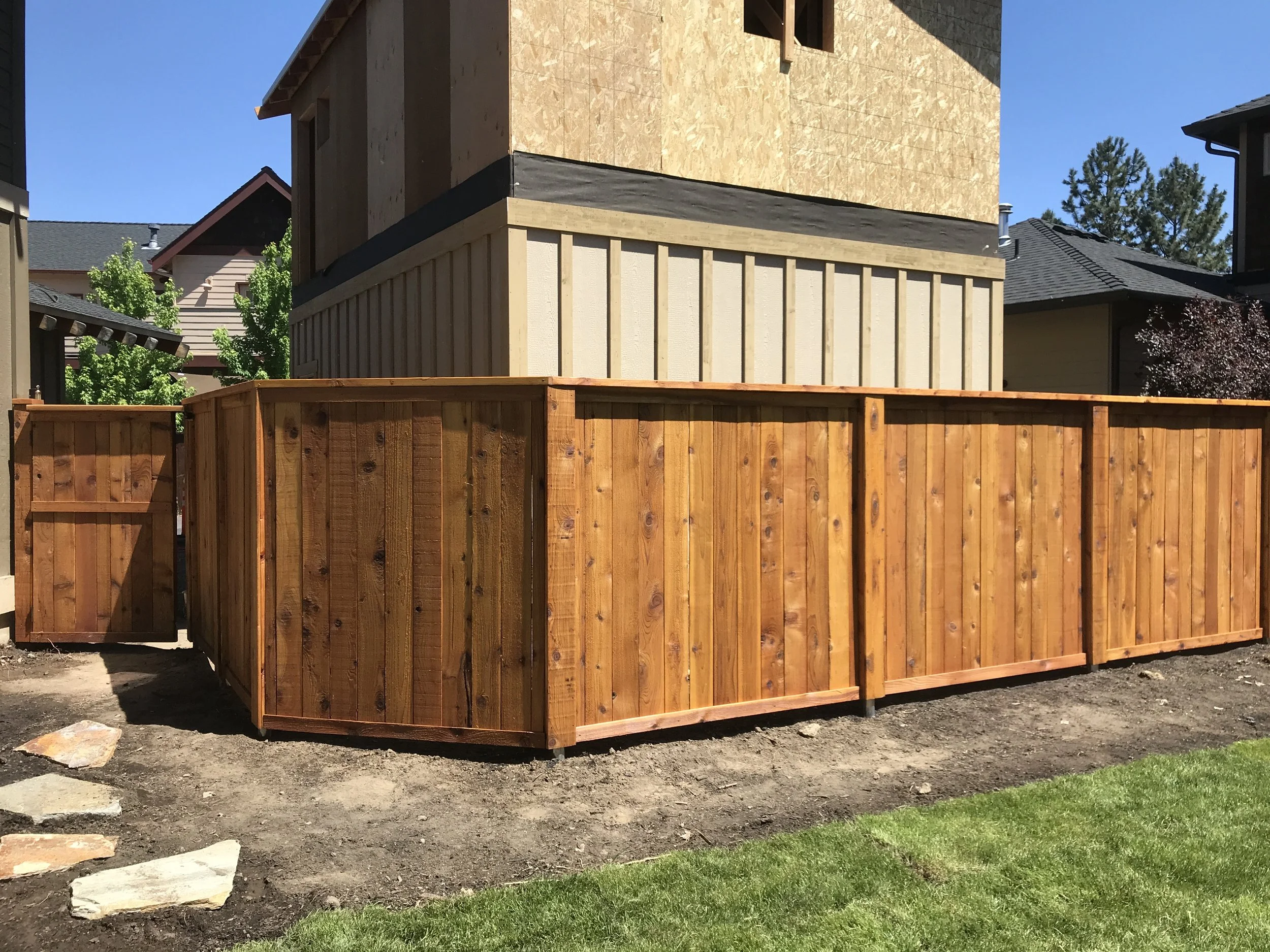 A wooden privacy fence surrounds a backyard area under construction, with a house in the background showing part of the structure and some trees, and a clear blue sky above.
