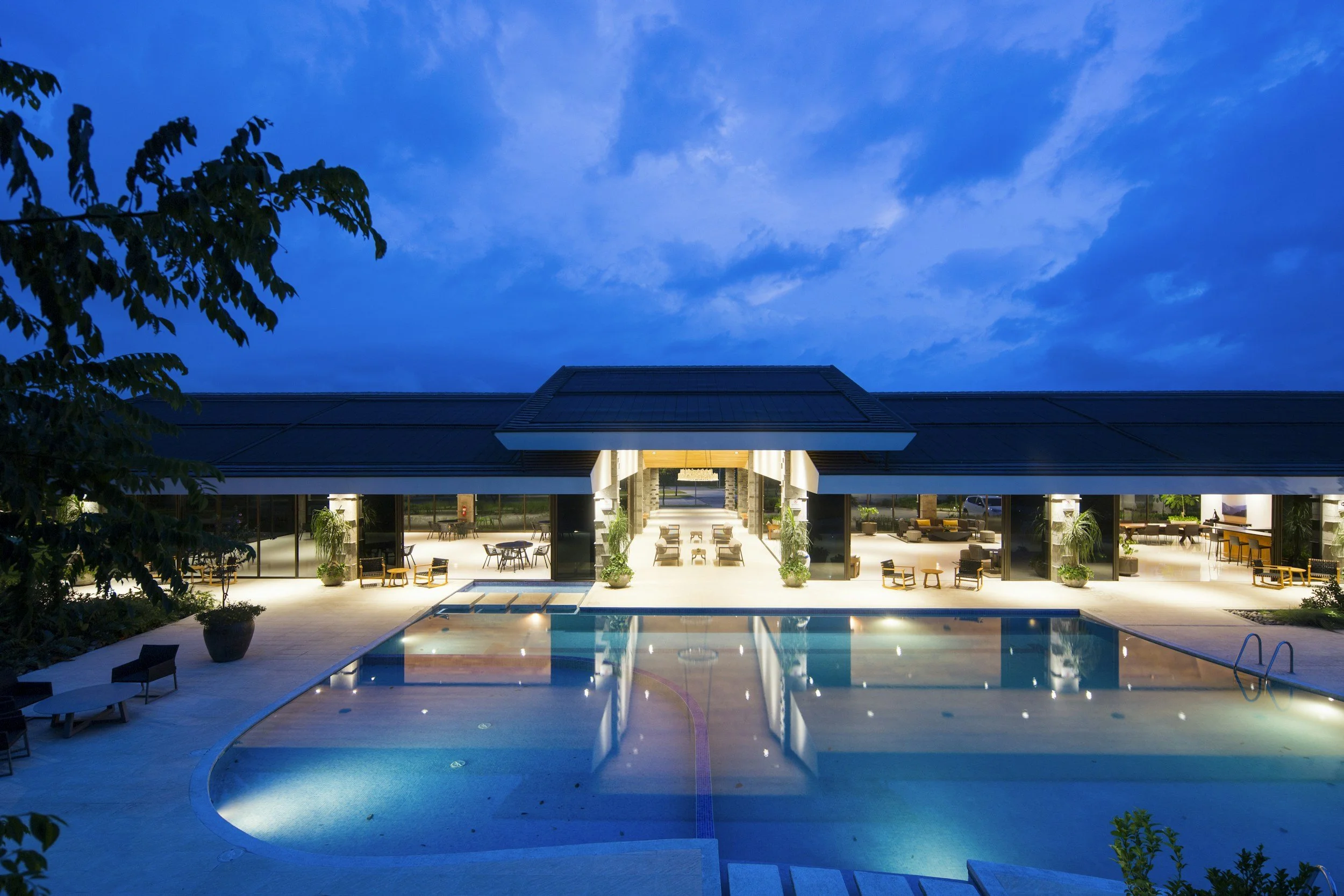 Luxury poolside lounge area at dusk with a modern building, outdoor seating, and blue-lit swimming pool.