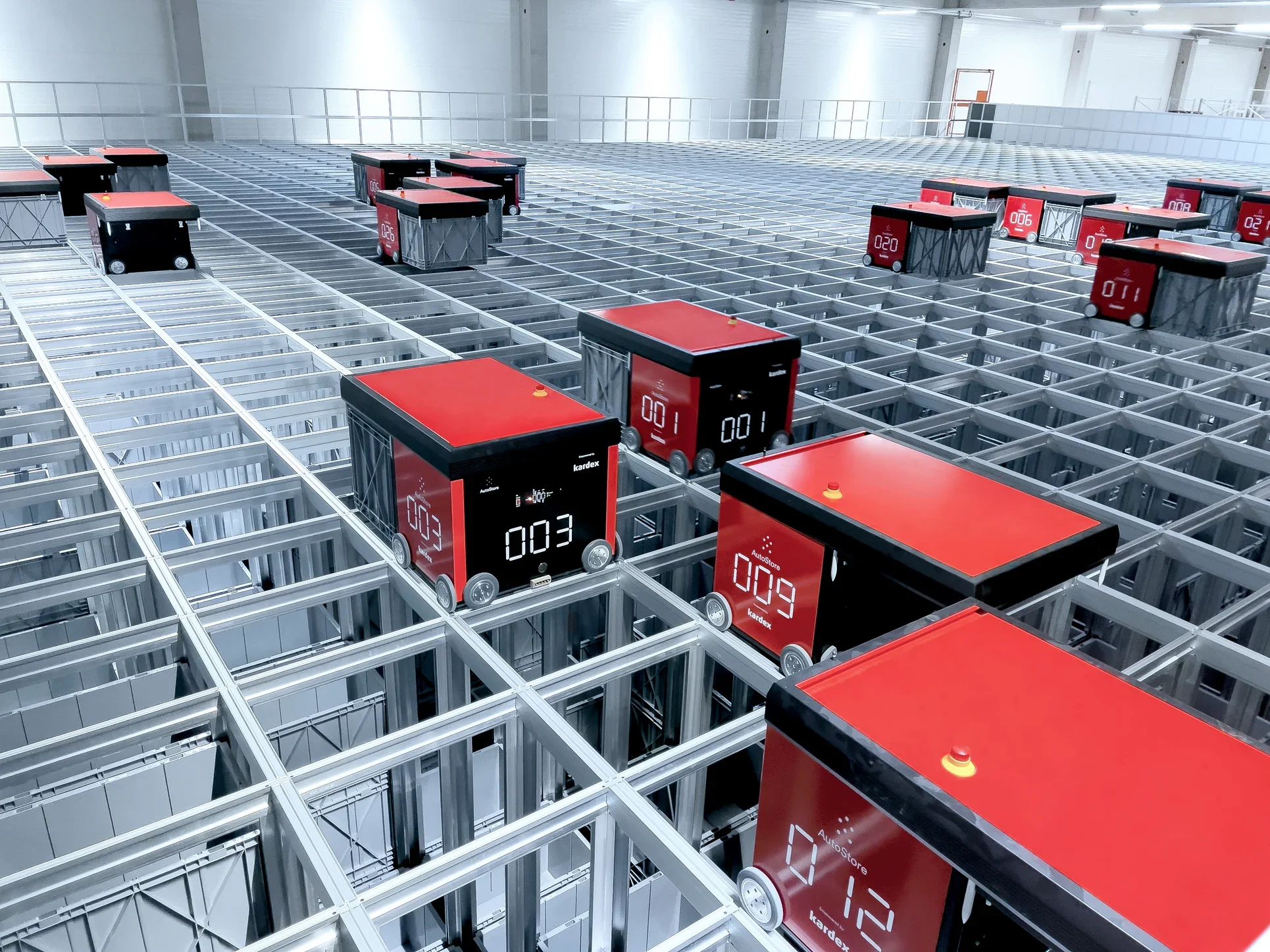 Multiple red and black autostore autonomous delivery robots on a metal grid structure in a warehouse.