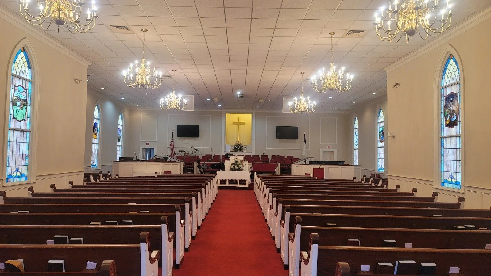 Interior of a church with rows of wooden pews, stained glass windows, chandeliers, and a raised stage with a cross and flags.