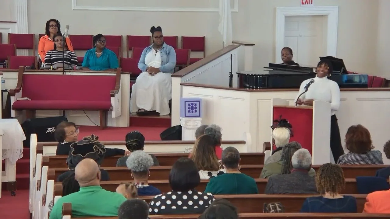 Woman speaking at a podium in church, with a pianist and several women seated on stage, and an audience of diverse people listening.