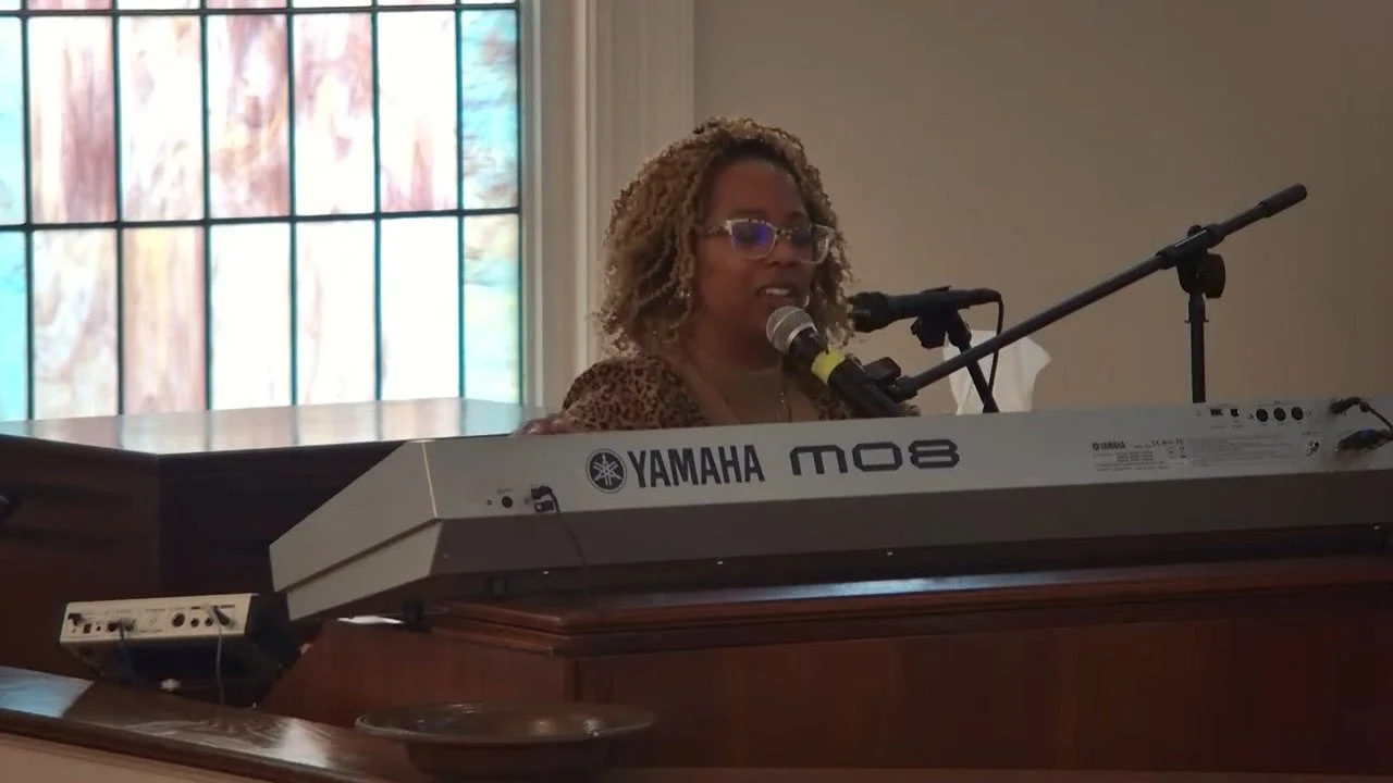 A woman with curly hair and glasses playing a Yamaha keyboard, speaking into a microphone in a room with a large window with stained glass.