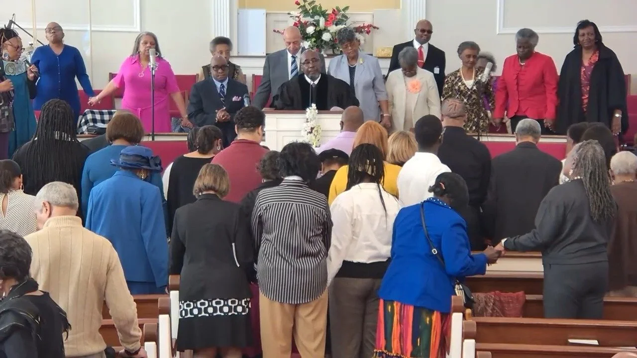 People gathered in church during a service, with some standing at the altar and others in the pews, a floral arrangement at the front.