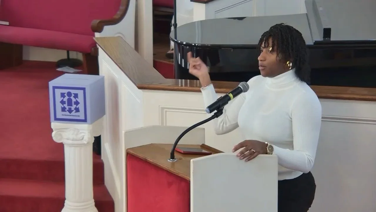 A woman with braided hair, wearing a white turtleneck and gold hoop earrings, is standing at a podium with a microphone, speaking in a church or similar setting.