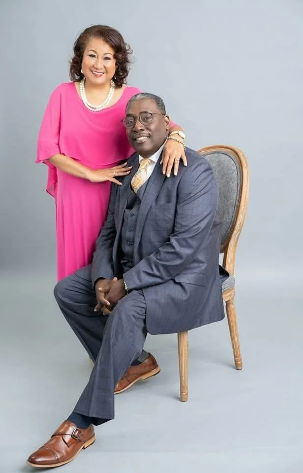 A woman in a pink dress and a pearl necklace standing beside a seated man in a gray suit, both smiling at the camera against a plain background.