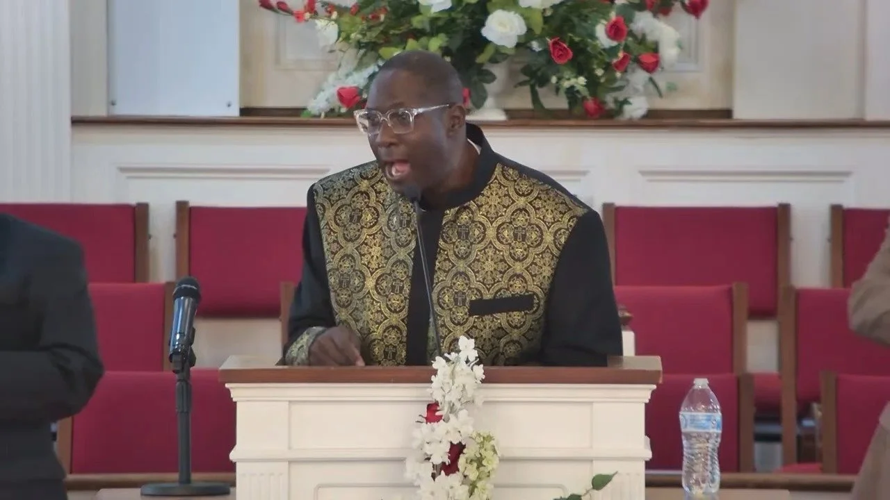 A man speaking at a church pulpit with a floral arrangement in the background, a microphone, and a water bottle in front of him.