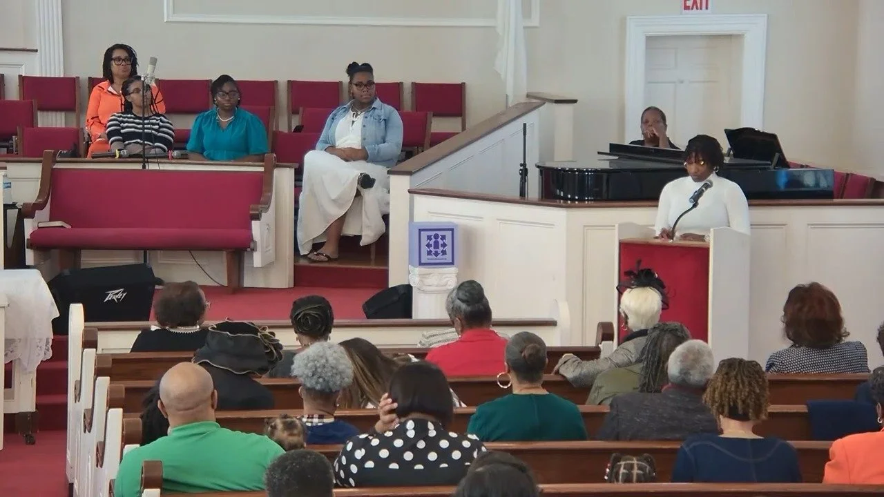 A woman stands at a podium giving a speech in a church surrounded by an audience, with a piano and several women seated on the elevated platform.