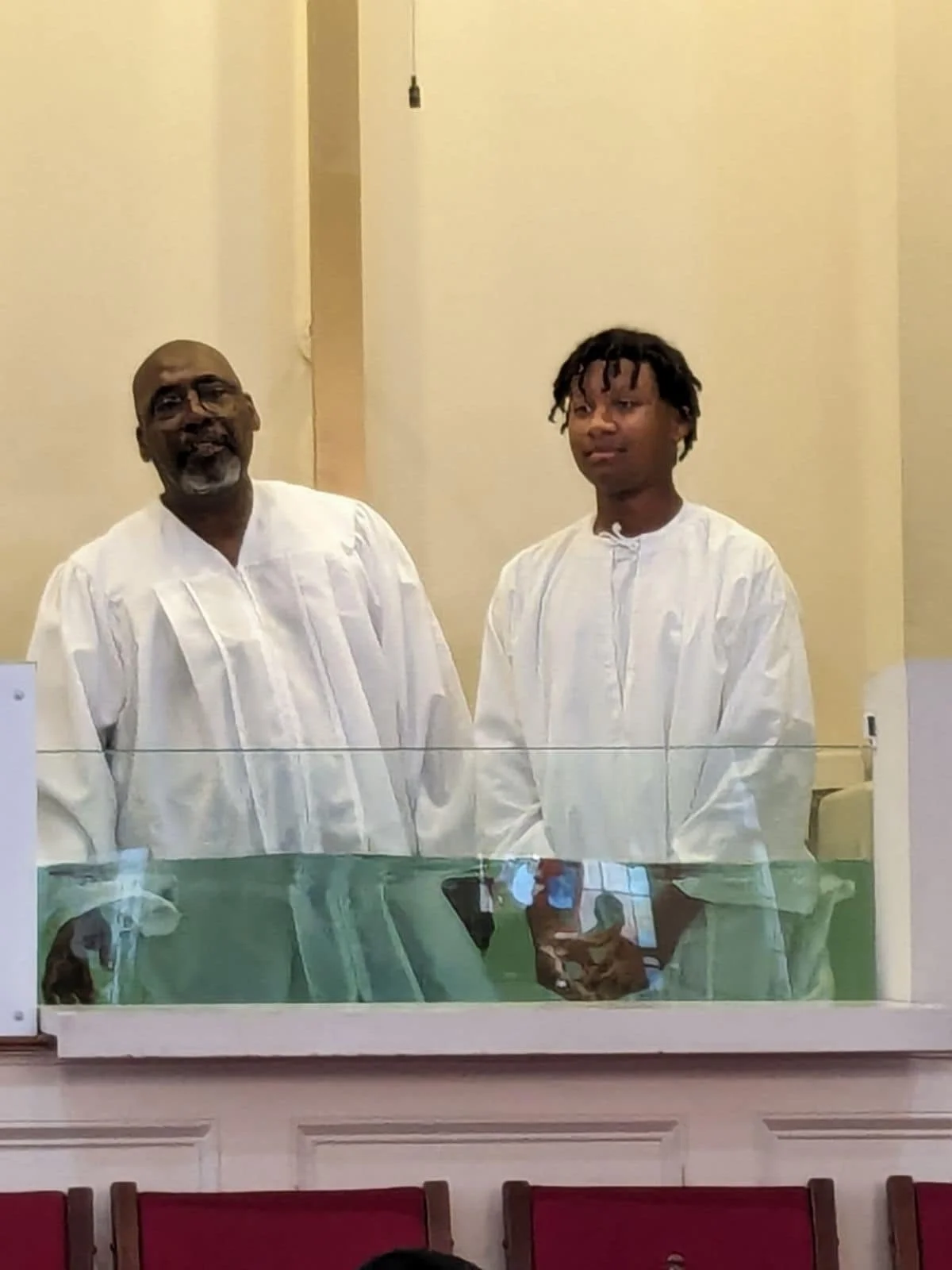 Two men wearing white robes standing behind a glass barrier, with reflections of electronic devices visible on the glass. They are in an indoor setting with cream-colored walls and red chairs visible in the foreground.