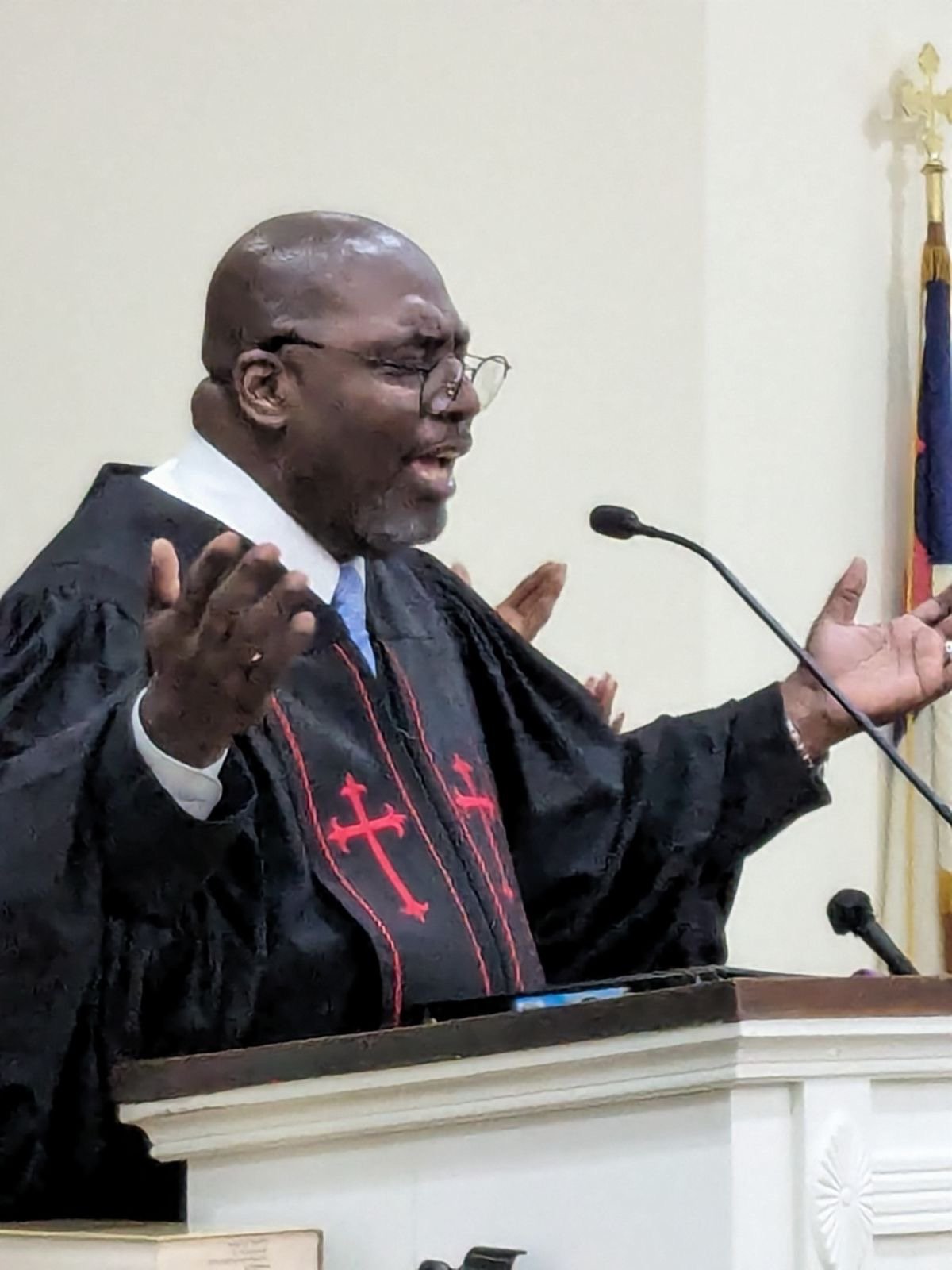 A man wearing a black robe with red crosses, standing at a pulpit with his eyes closed and hands raised in front of a microphone, in a church setting.