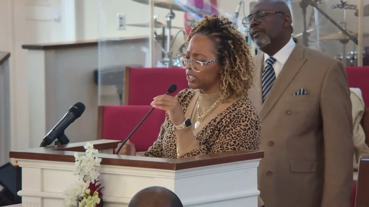 A woman with curly hair and glasses speaking at a podium with a microphone, wearing a leopard print top. A man in a brown suit with a striped tie stands behind her in a church or conference room setting.