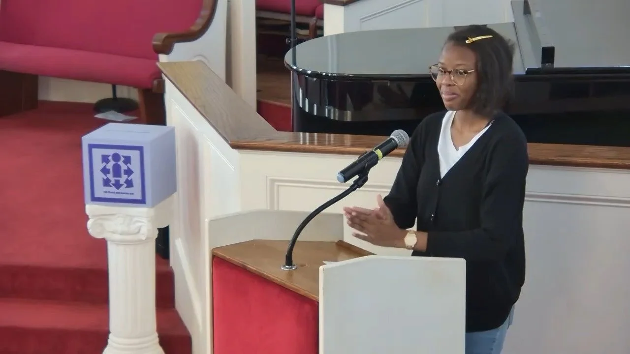 A woman with glasses, wearing a black cardigan and white shirt, standing at a wooden podium with a microphone in a church or auditorium. There are red upholstered pews and stairs in the background.