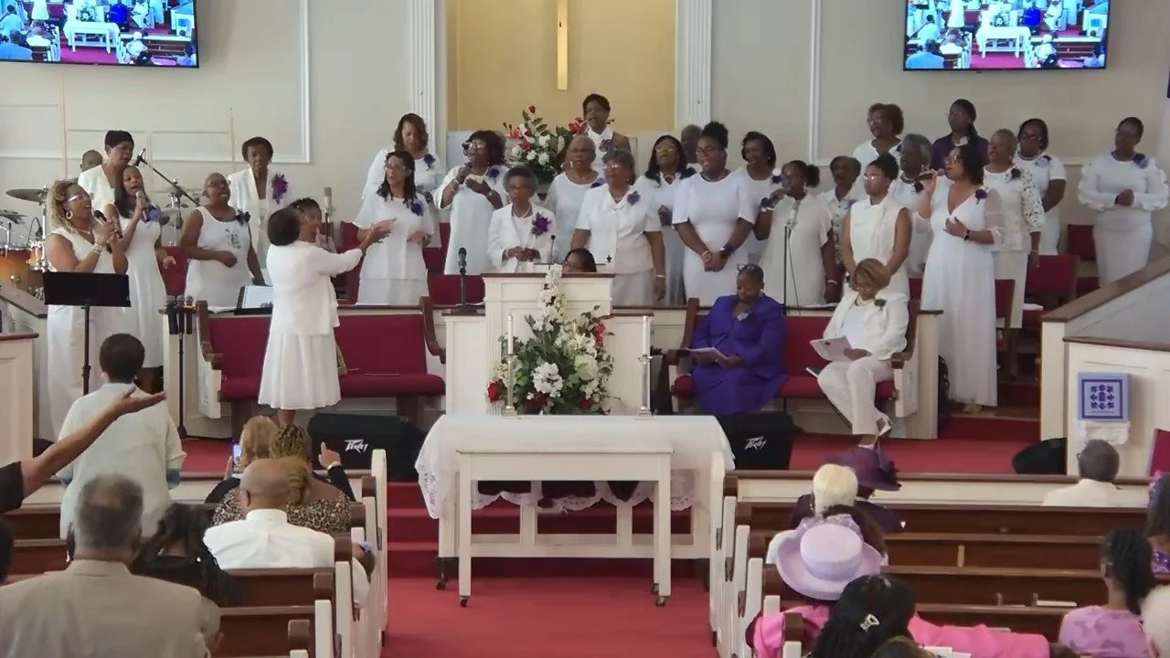 A church service with a choir dressed in white on the stage, some members holding microphones, and two women seated in the front row. The congregation is seated in pews, and the church is decorated with flowers.