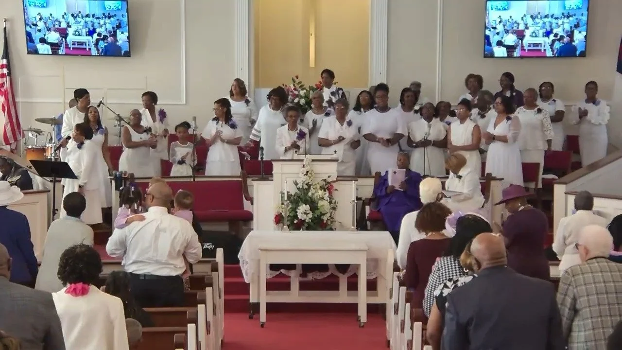 A church congregation gathered during a service, with a choir singing at the front, a woman in a purple gown seated near the pulpit, and people seated in pews facing the front. Two large screens show the congregation.