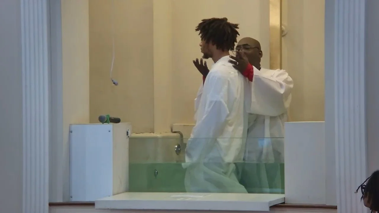 A man in white robes being baptized in a small indoor pool while a minister performs the baptism, with a microphone placed on the side of the pool.