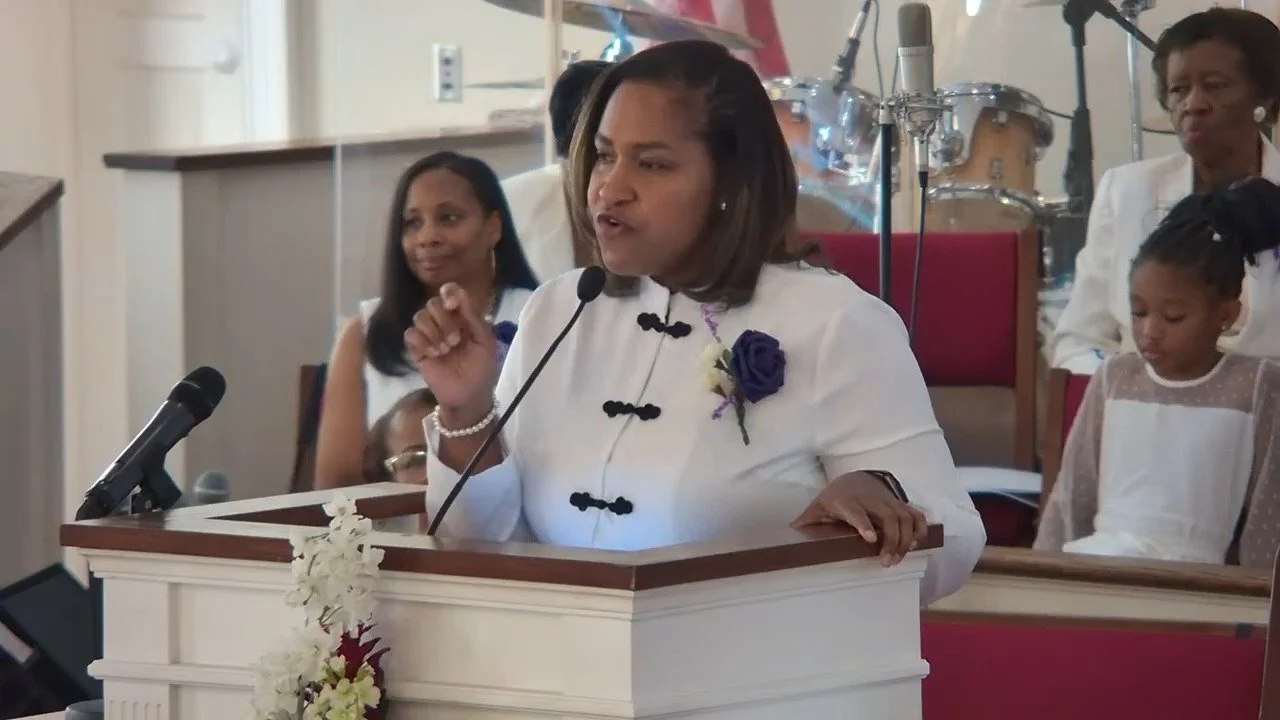 A woman speaking at a church podium during a service, wearing a white dress with a purple flower pin, and a microphone is attached to the podium. Behind her are women and a girl, some with jewelry, and musical instruments like drums, inside a church 