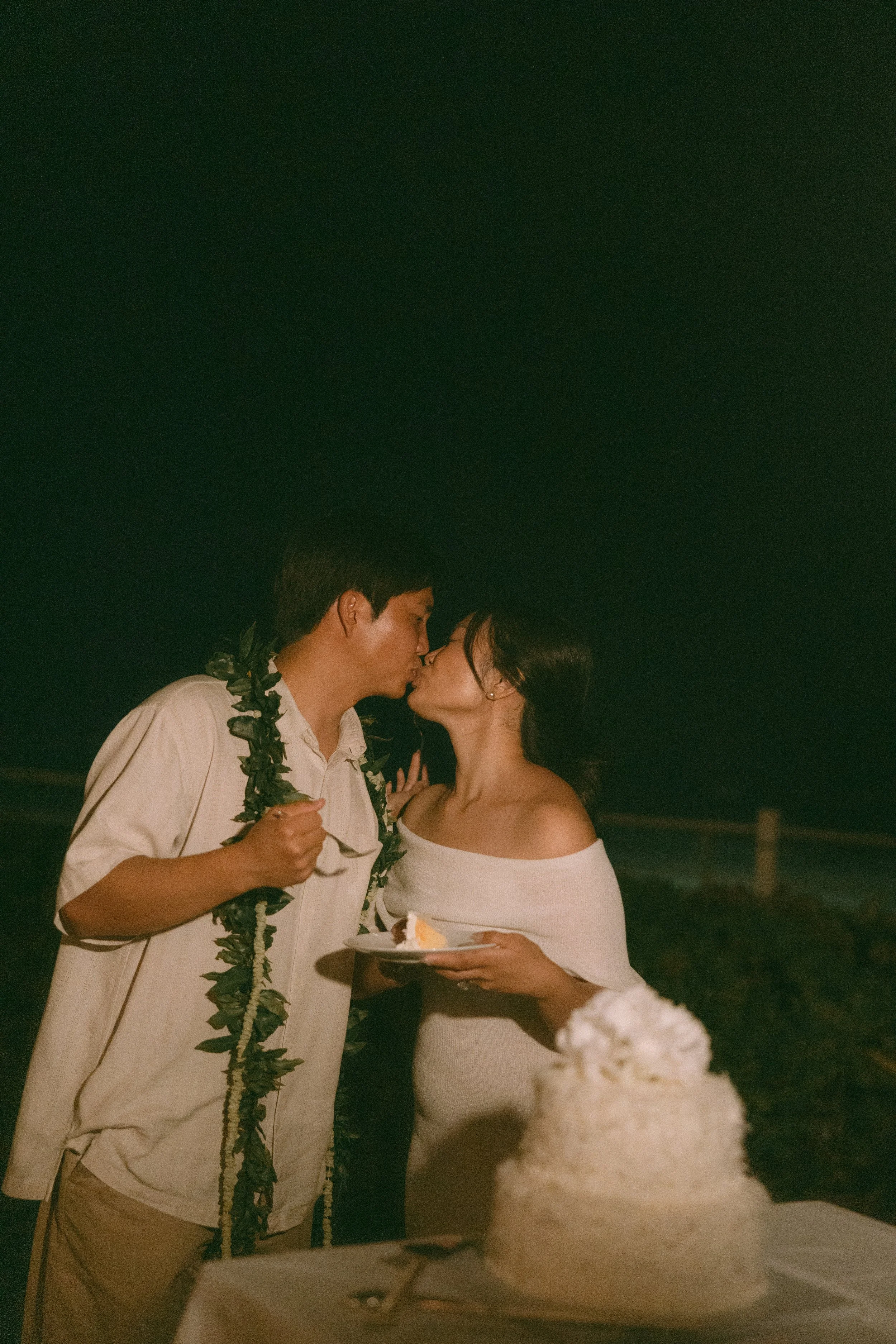 A couple sharing a kiss at night, with the man wearing a lei and a white shirt, and the woman holding a plate with a slice of cake, standing outdoors.
