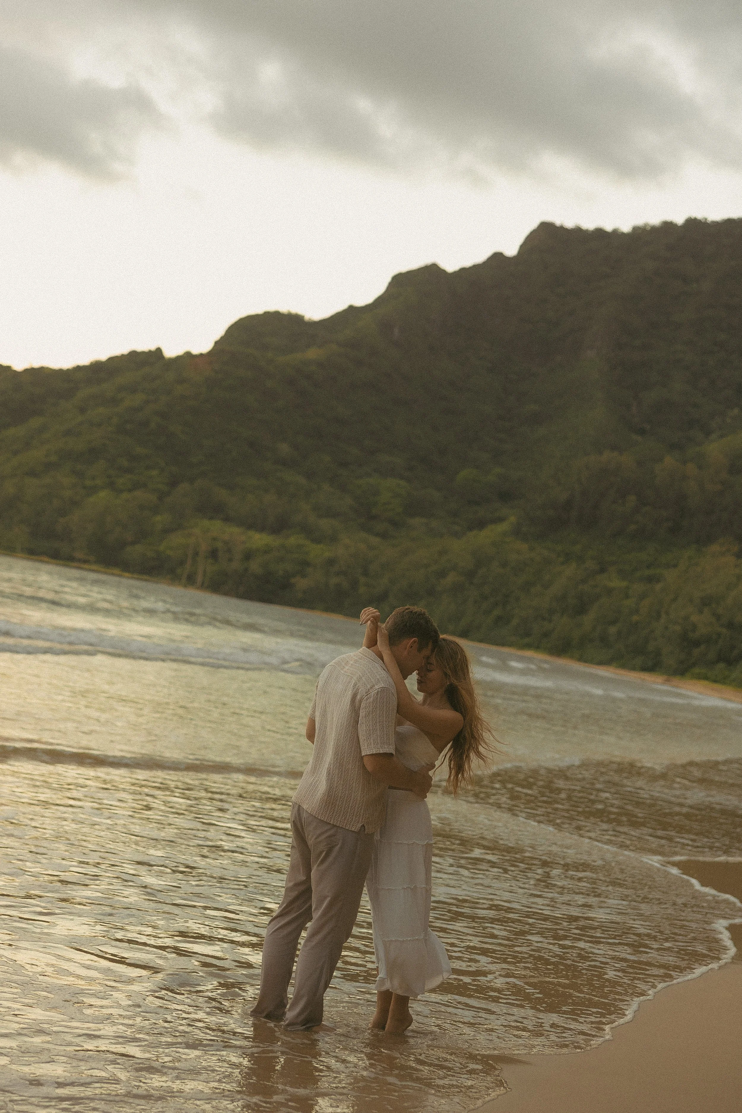 Couple sharing a forehead kiss at the shoreline on North Shore Oahu, dramatic mountain backdrop at golden hour, by Oahu couples photographer Maggie T Photography