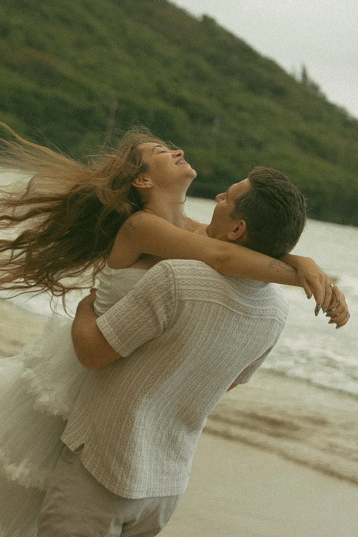Couple laughing and spinning on a windy North Shore Oahu beach, Ko'olau mountains in the background, candid couples photography by Maggie T Photography