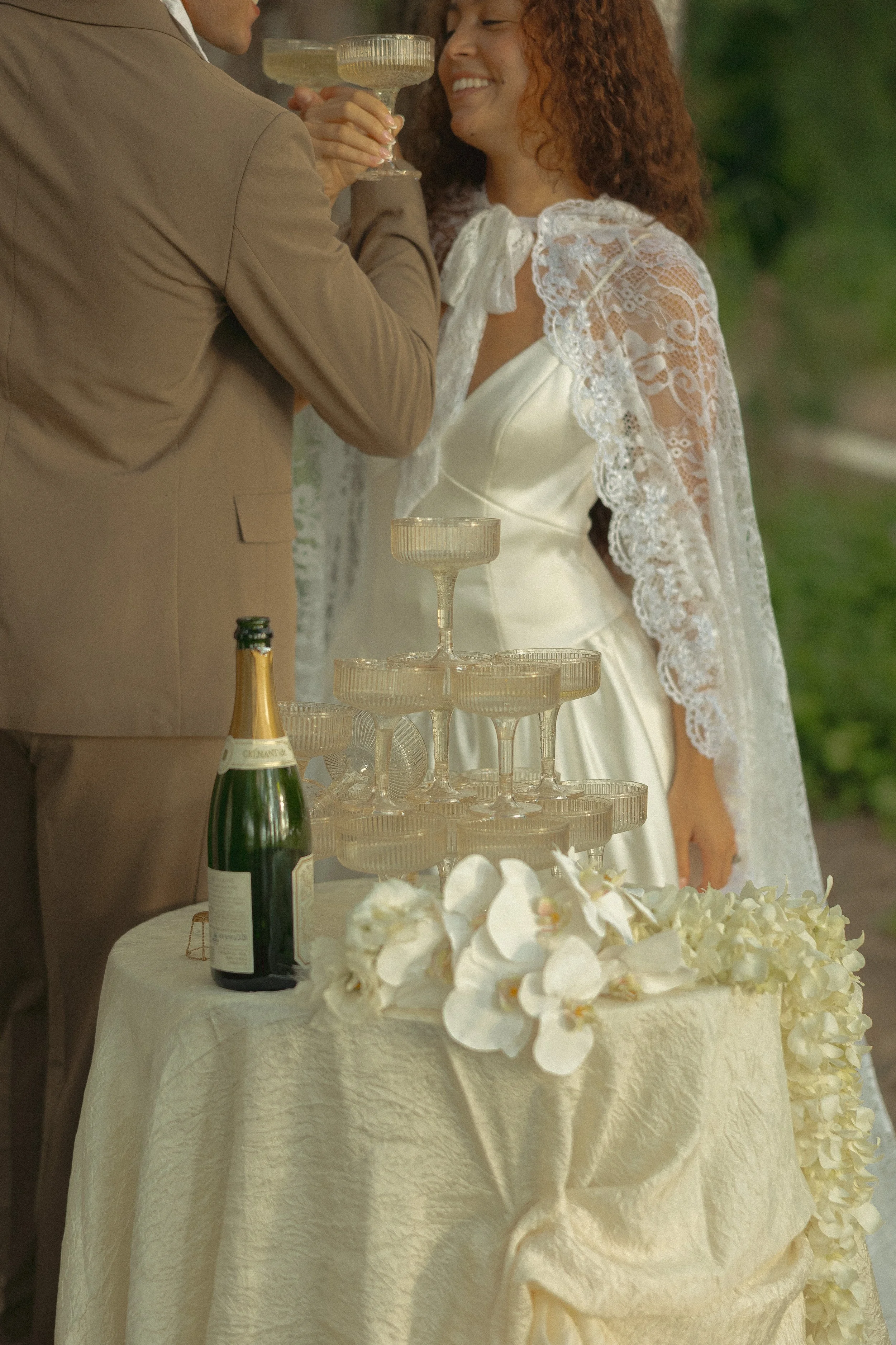 Bride and groom toasting with linked arms beside a champagne coupe tower and white orchids, Oahu elopement, film-toned
