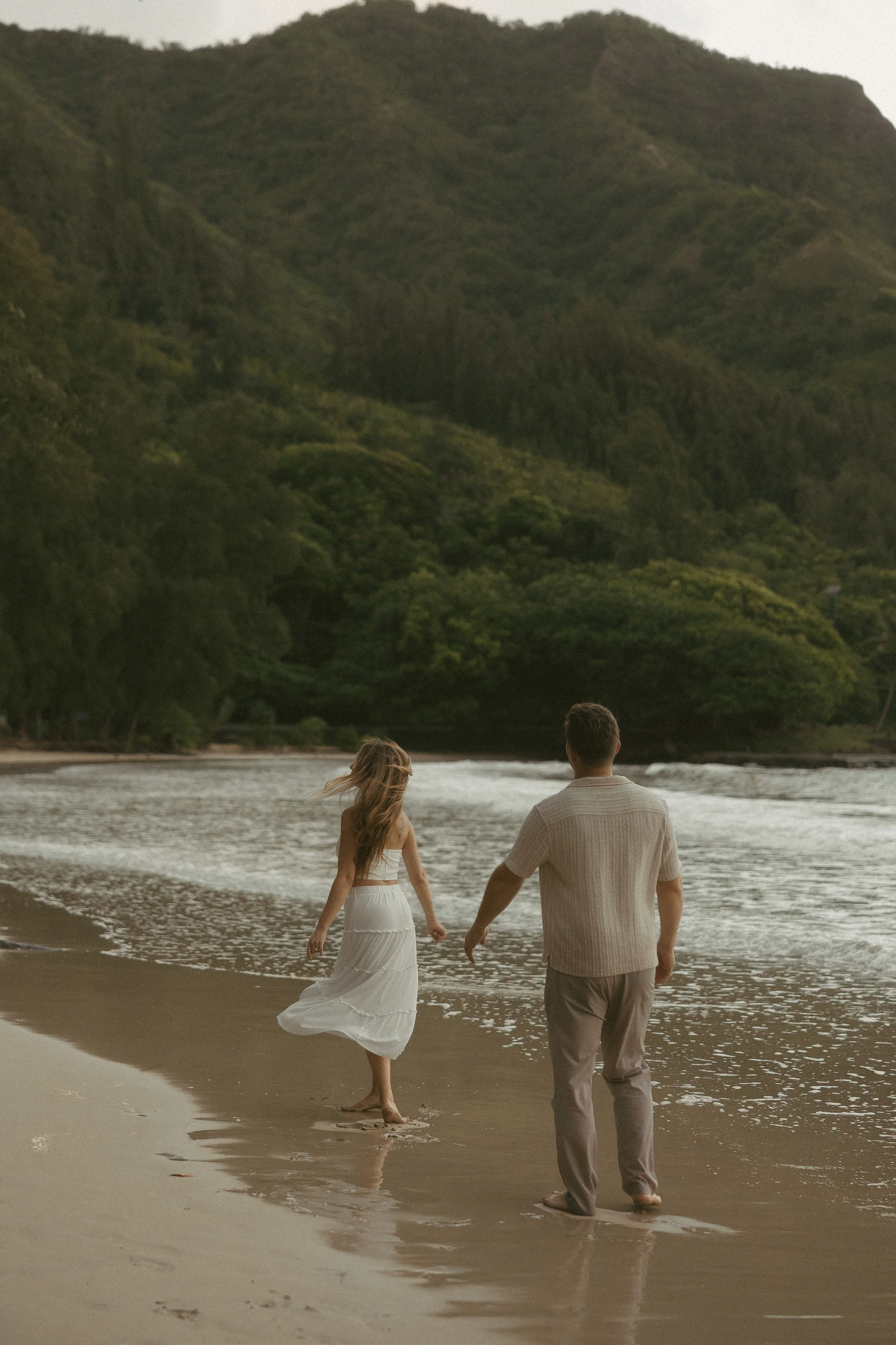 Couple walking barefoot along a windswept Oahu beach shoreline, lush green mountains in the background, shot by Oahu couples photographer Maggie T Photography