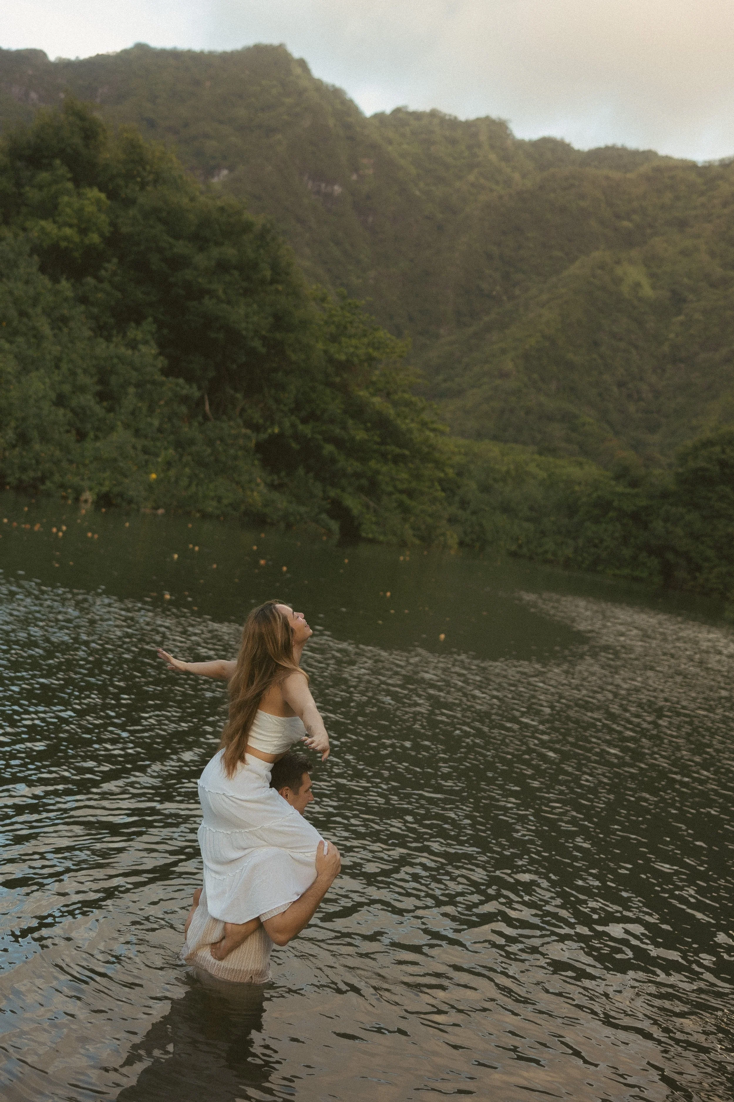 Man carrying woman on his back through a still mountain lake on Oahu's North Shore, lush green Ko'olau ridgeline behind them, by Oahu couples photographer Maggie T Photography